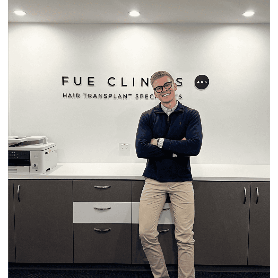 Smiling man in front of FUE Clinics sign. Hair transplant specialist with arms crossed. Clinic interior. FUE hair transplant expert.